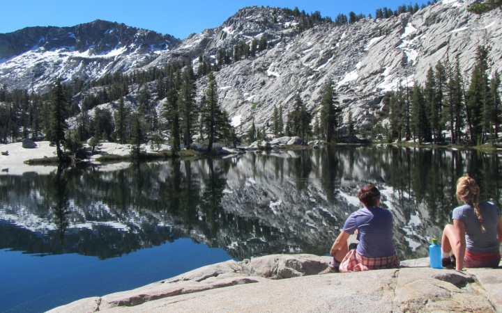 two students sit on a rock facing a blue alpine lake reflecting a mountainous landscape